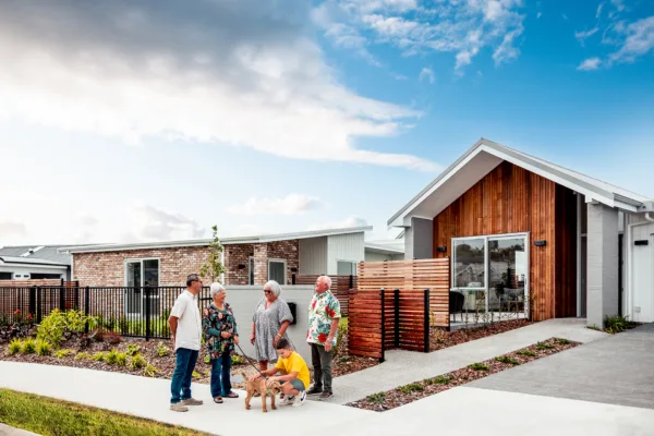 Photo of the front of one of the Vivid Living Homes. The house has attractive stained wood panelling on the entry wall and there is a wide footpath and a tidy front garden. There are two older couples chatting at the end of the driveway, as well as a child and a dog playing.