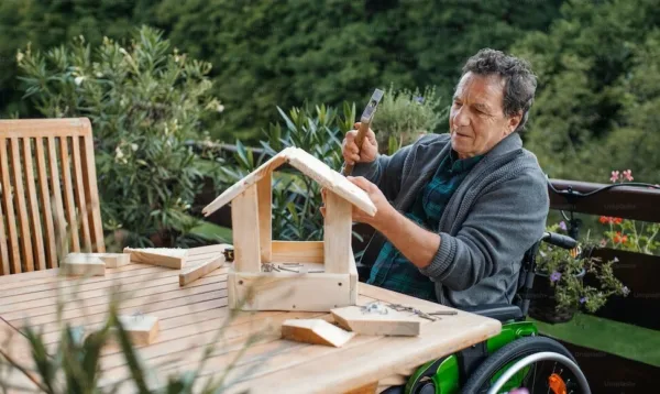 Photo of a man who uses a wheelchair on his deck building a wooden birdhouse.