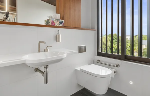 A bathroom with a wall-mounted sink and toilet, a large mirror and a wide window with vertical slats for privacy.