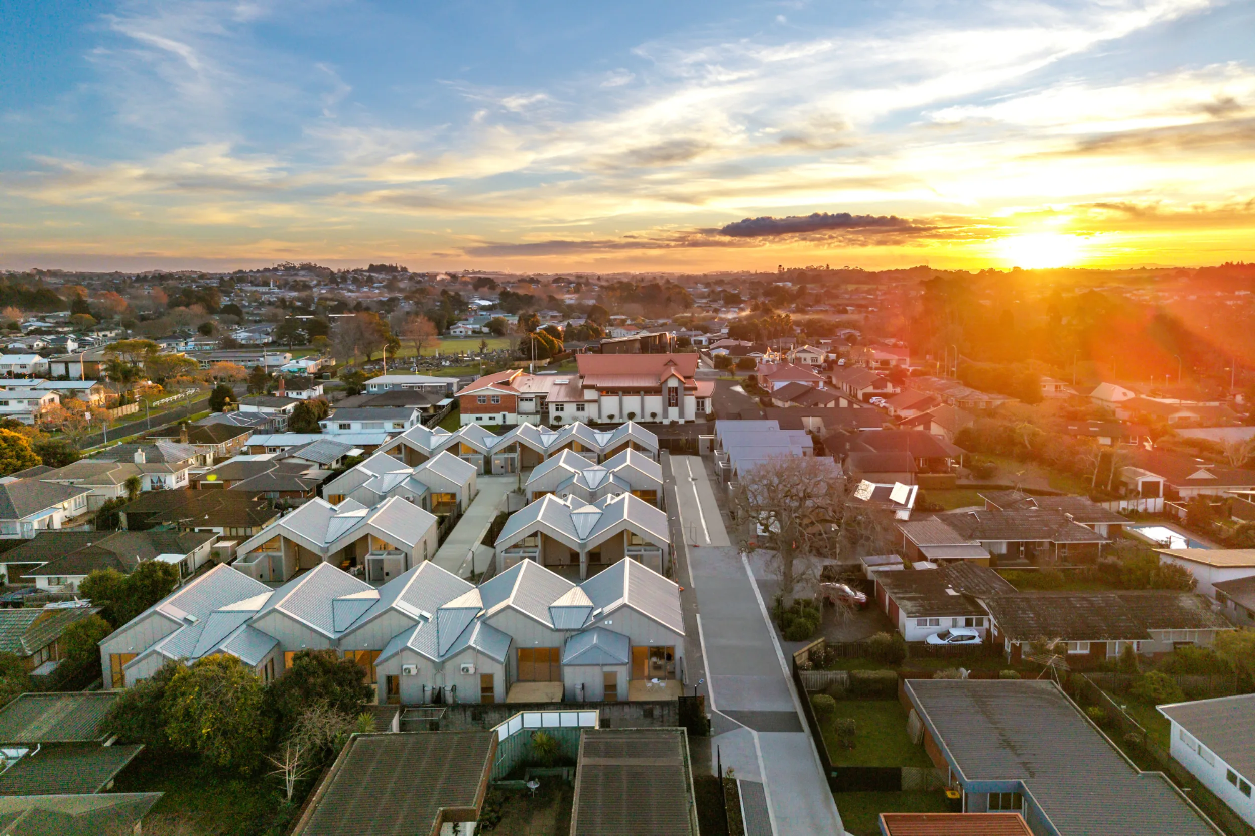 Aerial view of Notre Dame Living in Pukekohe at sunset. Rows of single-level homes with pale grey cladding and distinctive pitched metal roofs are clustered in a village-style layout, with paved paths running between them and a wide driveway along one side. The development sits among established suburban houses and trees, with a large brick church building behind it and the sun setting over the town on the horizon.