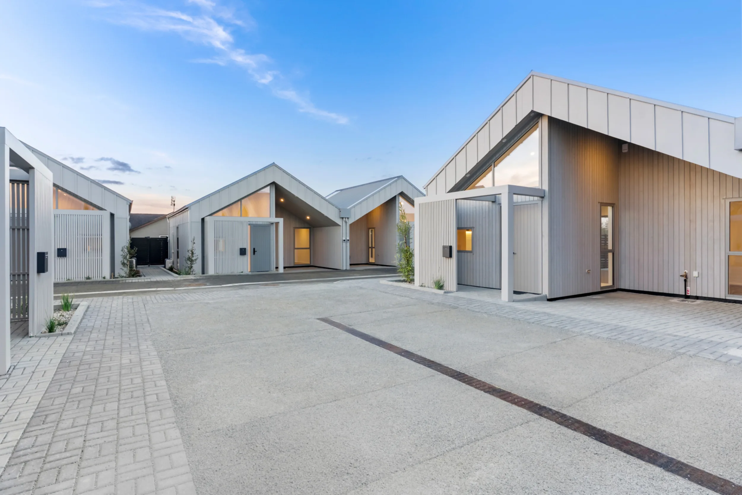 Exterior of several Notre Dame Living homes at dusk, with pale grey vertical cladding and steep pitched gables. Each home has a covered entry with a slatted screen and letterbox, and warm light glows from the large windows. A wide, flat paved courtyard with brick-patterned edging and a drainage channel leads to the front doors, with young plantings along the edges.