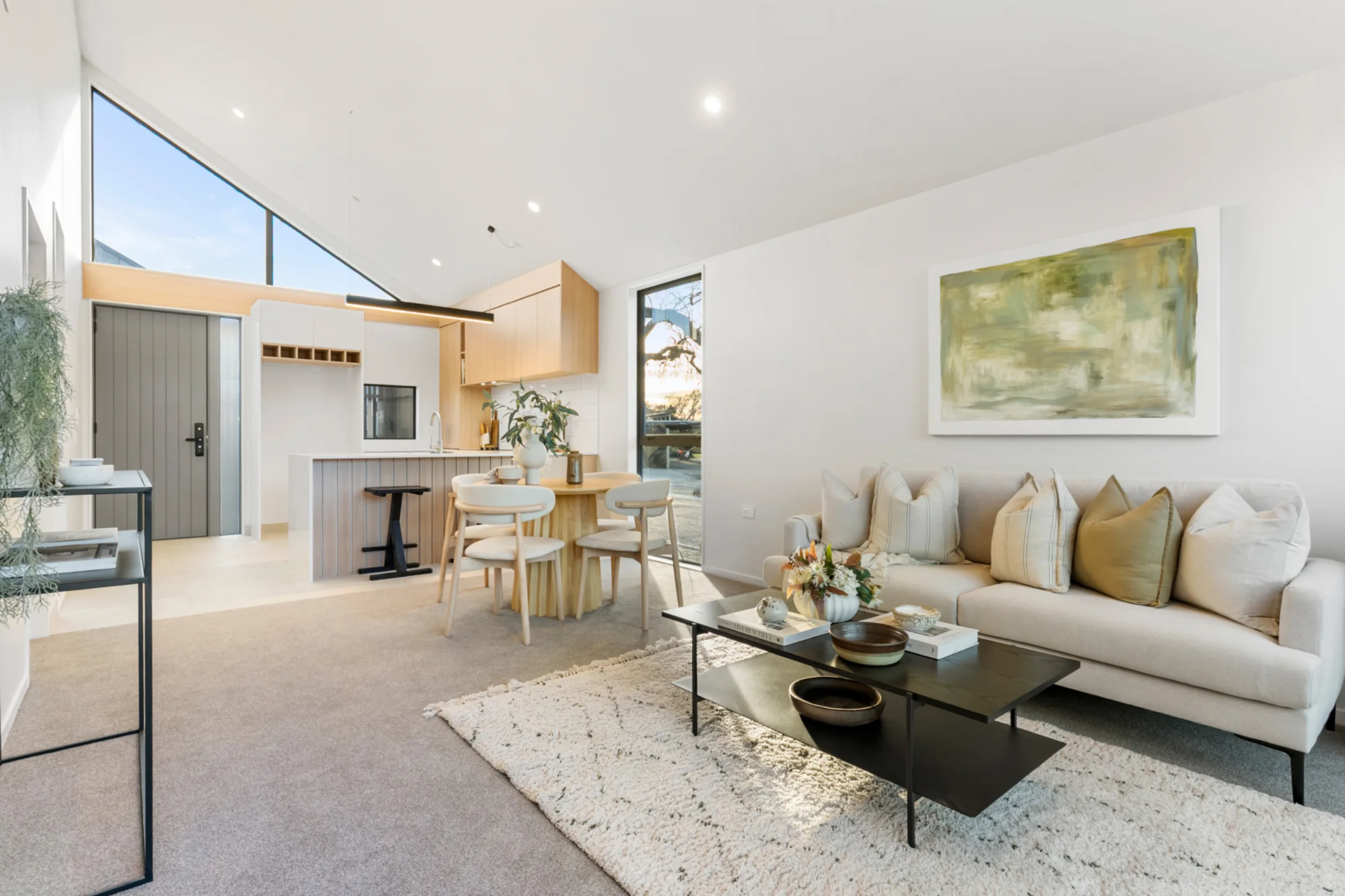 Bright open-plan living, dining and kitchen area in a Notre Dame Living home. A cream sofa with cushions faces a black coffee table on a white rug, with a large green abstract painting on the wall above. Beyond, a round timber dining table with four chairs sits in front of a kitchen with oak-look cabinetry and a vertical-slatted island. A high triangular window under the raked ceiling fills the room with natural light, and the front door is visible at the far end.