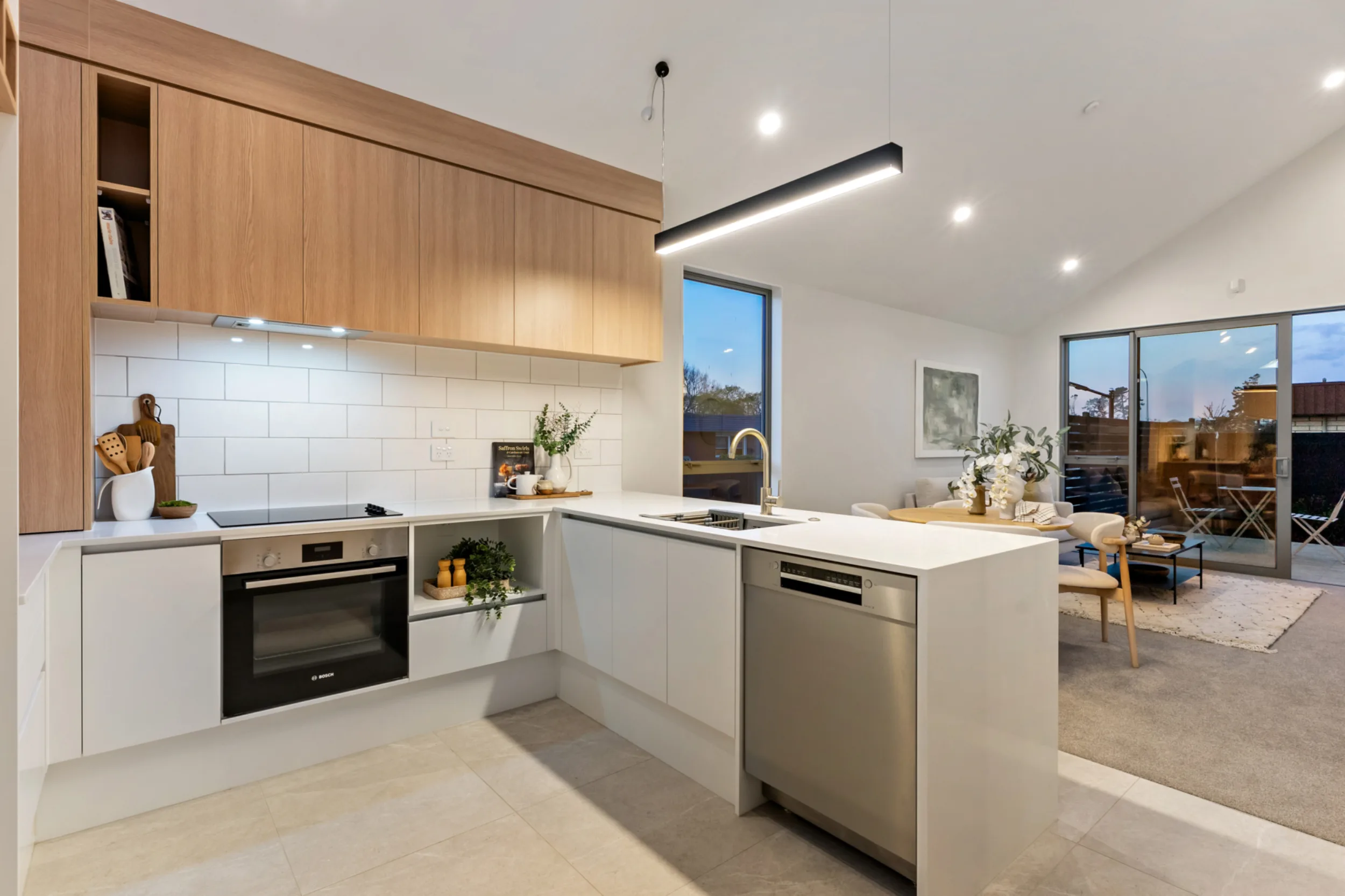 Kitchen in a Notre Dame Living home, with white lower cabinets, oak-look upper cabinets and a white subway-tile splashback. The L-shaped layout includes a wall oven, induction cooktop, dishwasher and a brass gooseneck tap at the sink. A linear pendant light hangs from the raked ceiling. The kitchen opens onto a dining and living area with sliding glass doors leading to an outdoor patio.