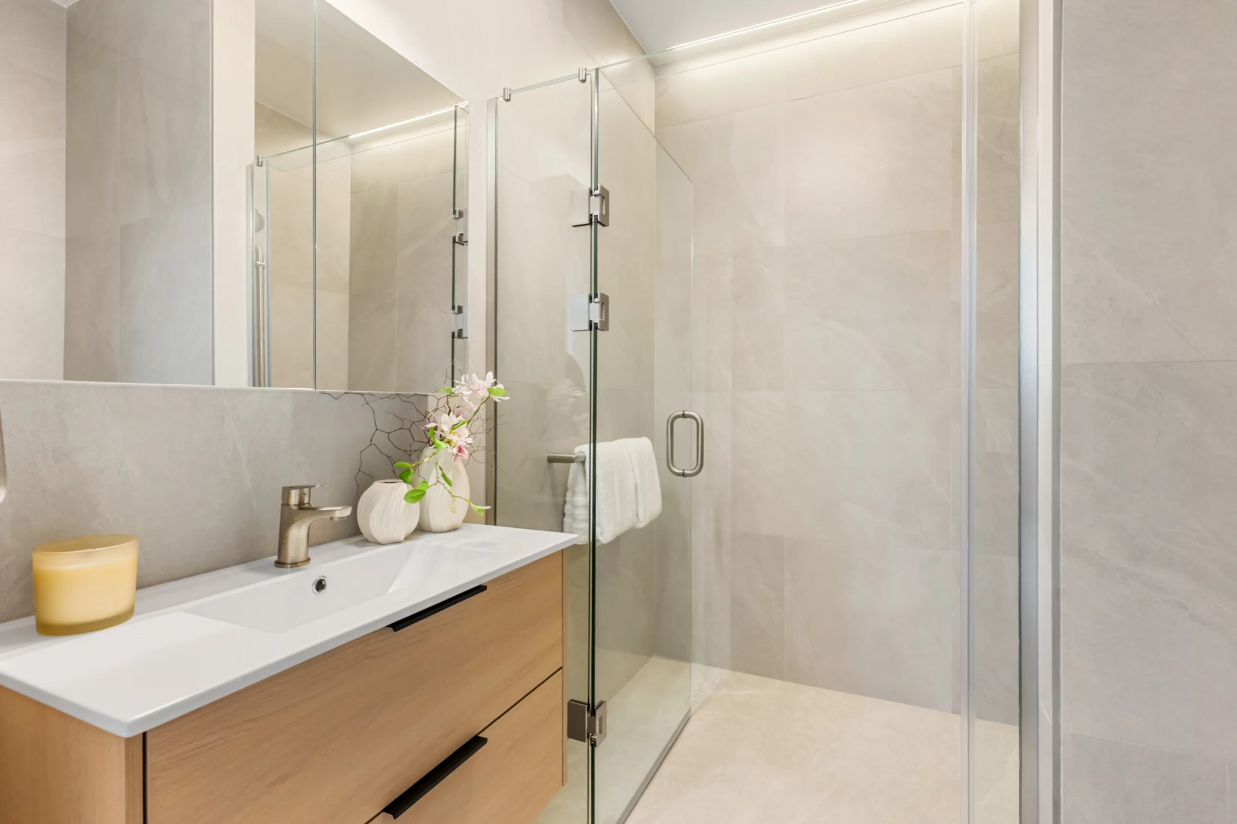 Bathroom in a Notre Dame Living home, finished in large pale grey stone-look tiles. A wall-hung timber vanity with a white basin and brushed nickel mixer tap sits beneath a wide mirrored cabinet. Beside it is a spacious shower with a frameless glass door and a level, step-free floor that continues the bathroom tiling. A white towel hangs on a rail inside the shower.