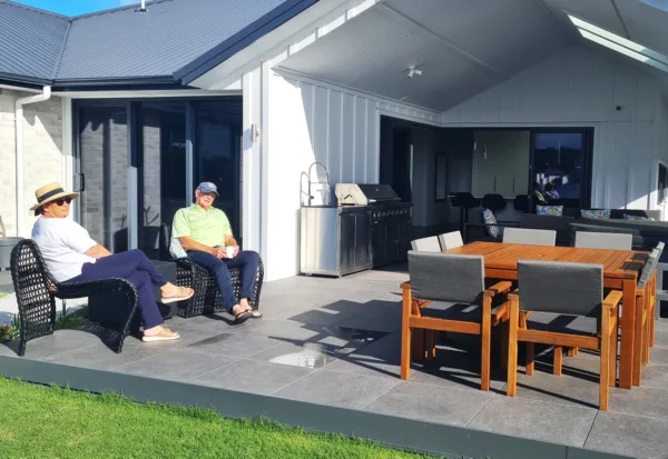 Raewyn and Pete enjoying their outdoor patio in front of their home. There is a table and chairs, and the ground is concrete. The home is white, with large sunny windows and a dark grey roof.