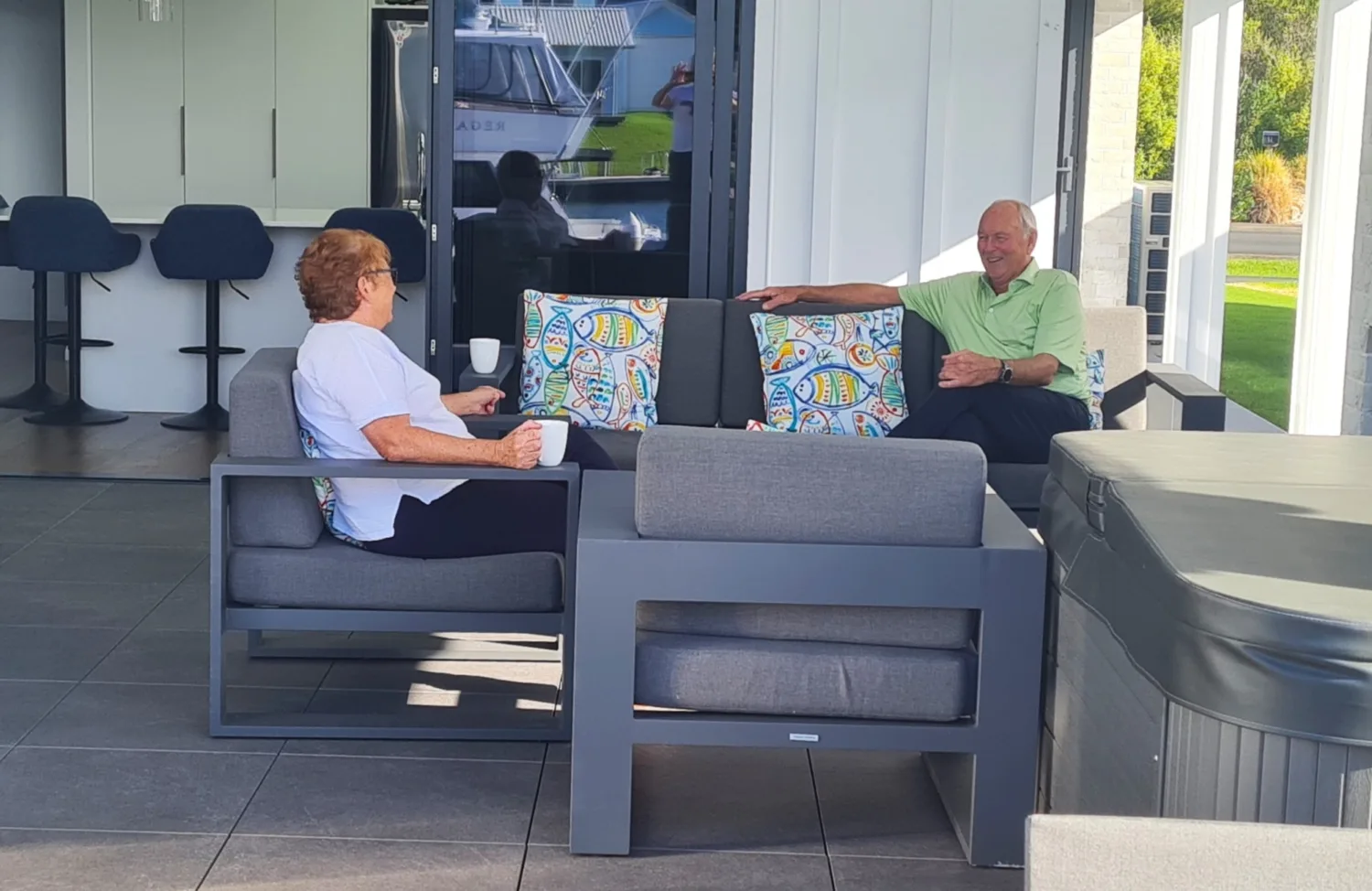 Raewyn and Pete are sitting in their patio area on outdoor couches. There is a spa pool on the right, a barbeque on the left and behind them is the french doors into the kitchen.